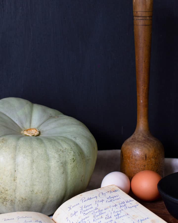 A moody kitchen vignette including a pumpkin, recipe book, eggs and a sauerkraut pounder