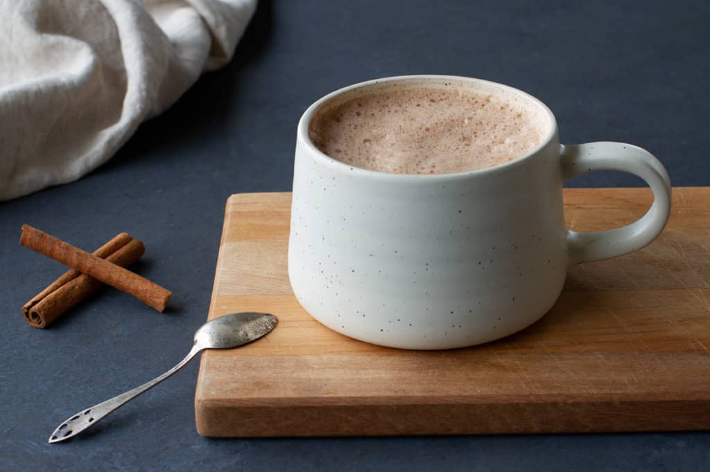 A cream mug of hot chocolate in a cream cup on a wooden board following my hot cacao recipe