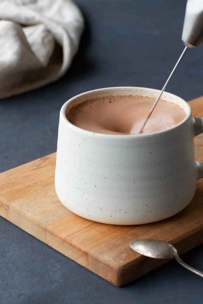 A cream mug of hot chocolate being whisked on a wooden board following my hot cacao recipe