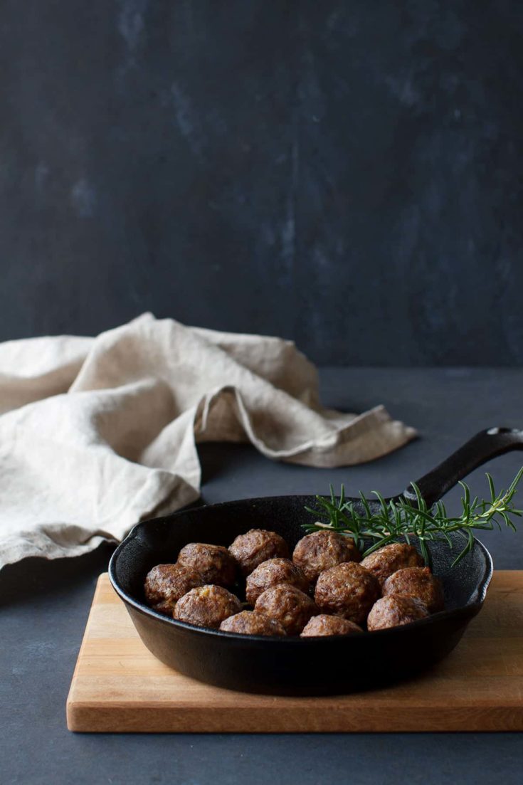 Meatballs without eggs alongside a sprig of rosemary in a cast iron pan which is placed on a wooden board. A linen tea towel is in the background.