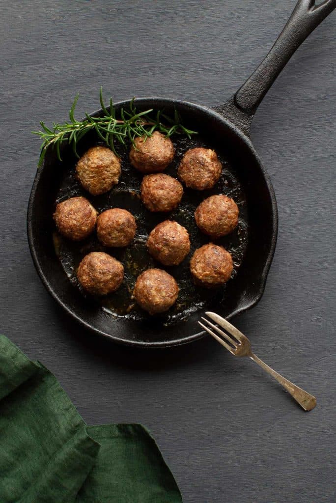 A flatlay of meatballs without eggs alongside a sprig of rosemary in a cast iron pan. A green tea towel is to the side. Moody scene.