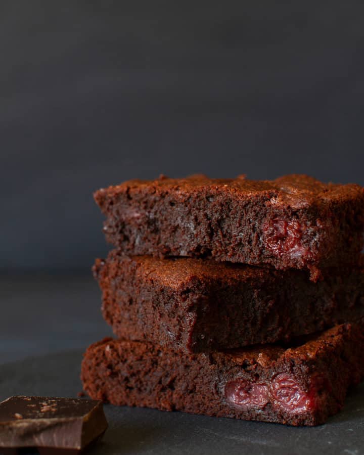 Three stacked squares of cherry brownies on a shingle next to a square of dark chocolate. Dark background.