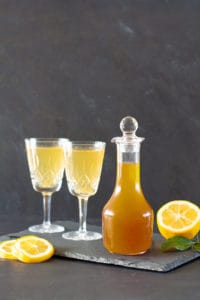 A glass jar of lemon cordial next to two vintage wine glasses with lemonade on a black shingle tile.
