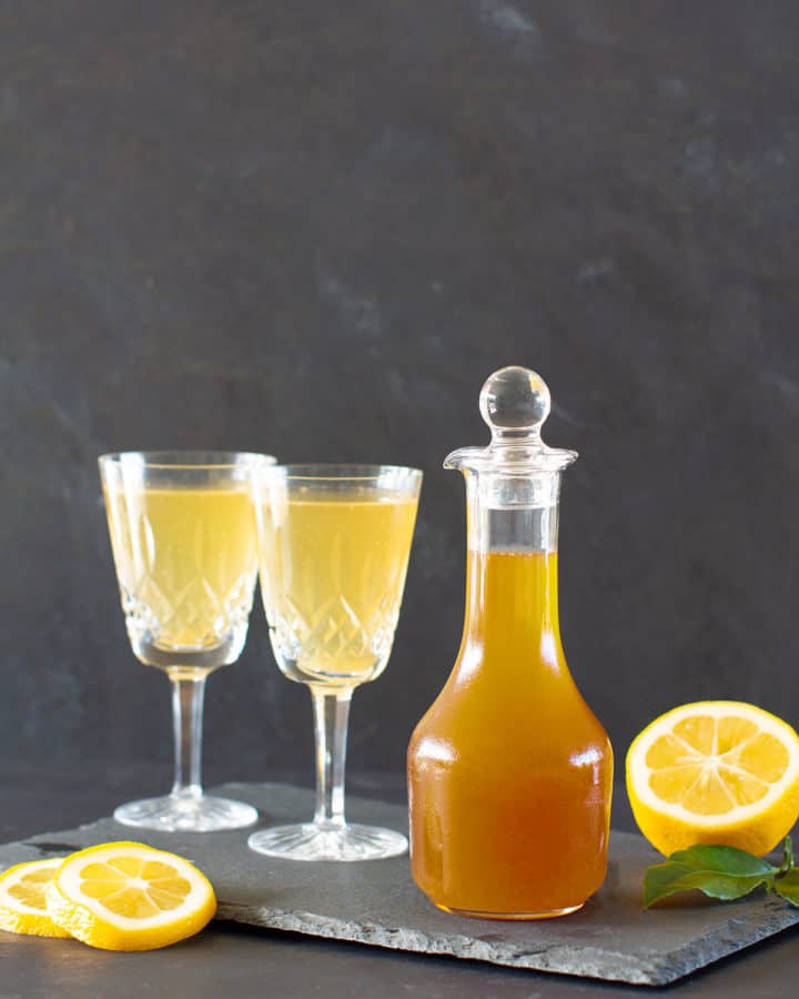 A glass jar of lemon cordial next to two vintage wine glasses with lemonade on a black shingle tile.