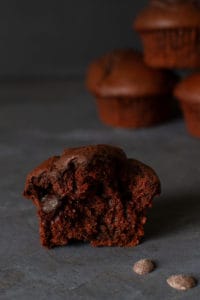 Close-up of a half triple chocolate muffin next to some chocolate drops and a stack of blurred muffins in the back.
