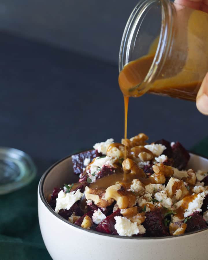 A bowl of roasted beetroot walnut and feta salad on a green tea towel. A jar of dressing is being poured onto the salad.