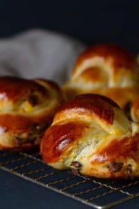 Close-up of a chocolate knotted bun on a cookie rack. A couple of buns and a linen tea towel are blurred out in the background.