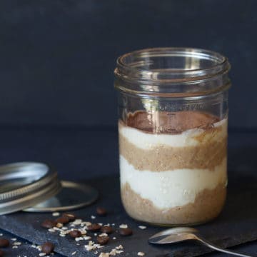 A clear jar of tiramisu overnight oats on a shingle plate. Mason jar lid, spoon, oats, and coffee beans are next to it.