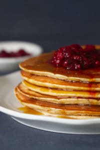 A stack of whey pancakes with raspberries and maple syrupon a white plate next to a small bowl of raspberries and a tea towel.