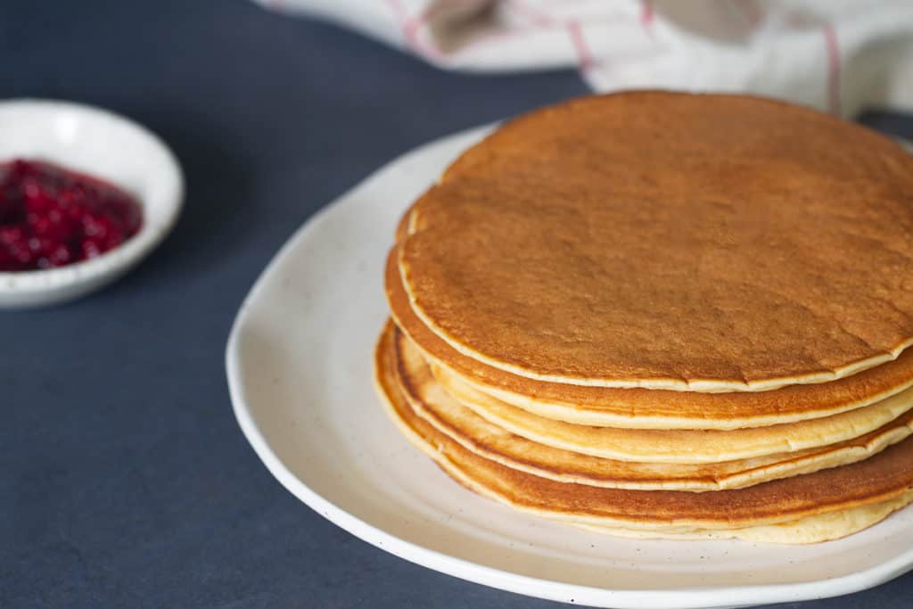 A stack of whey pancakes on a white plate next to a small bowl of raspberries and a tea towel.
