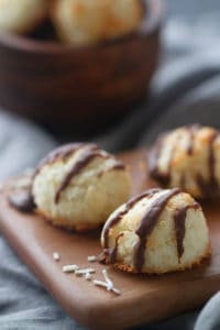 Three coconut macaroons (Kokosmakronen) on a wooden board and grey tea towel. A bowl of extra macaroons is blurred out in the background.